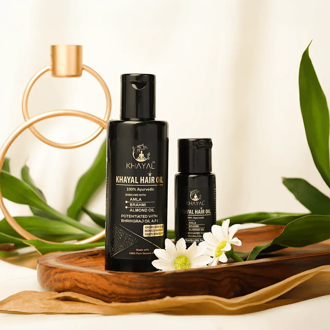 Two black bottles of Khayal Hair Oil are placed on a wooden tray with white flowers and green leaves around them.