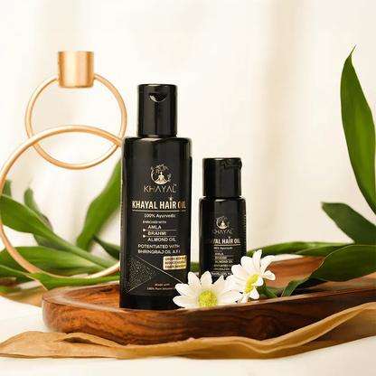 Two black bottles of Khayal Hair Oil are placed on a wooden tray with white flowers and green leaves around them.